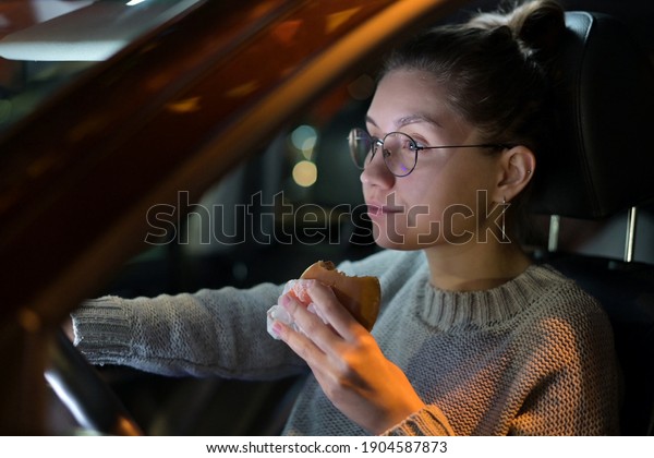 young woman\
eating a burger while sitting in her car in a parking lot in the\
evening. Busy schedule, food on the\
go