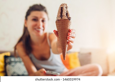 Young Woman Eating Chocolate Ice-cream In Cone Sitting On Couch At Home. Girl Holding And Showing Dessert