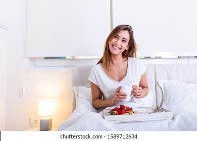 Young woman eating healthy breakfast in bed. Young woman eating breakfast in bed drinking coffee. Beautiful young woman smiling and eating strawberries .