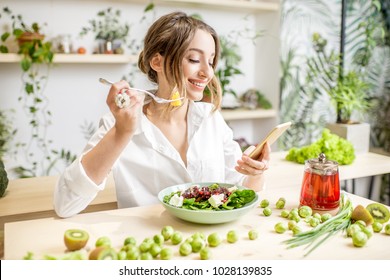 Young woman eating healthy food sitting with smartphone in the beautiful interior with green flowers on the background - Powered by Shutterstock