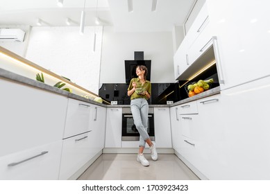 Young Woman Eating Healthy Salad On The Kitchen At Home, Wide Angle View On The Modern White Interior