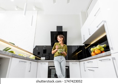 Young Woman Eating Healthy Salad On The Kitchen At Home, Wide Angle View On The Modern White Interior