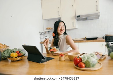 A young woman eating a healthy salad and working on a tablet in a modern kitchen surrounded by fresh vegetables. - Powered by Shutterstock