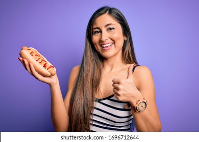 Young woman eating hotdog with ketchup and mustard over purple background happy with big smile doing ok sign, thumb up with fingers, excellent sign