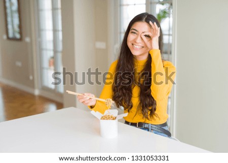 Young woman eating noodles in delivery box with happy face smiling doing ok sign with hand on eye looking through fingers