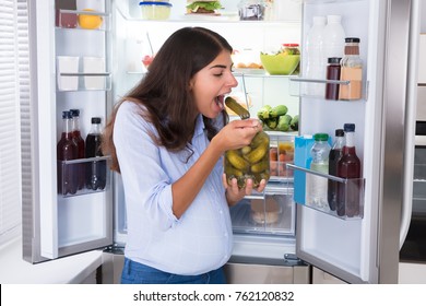 Young Woman Eating Pickle From Jar Near Refrigerator