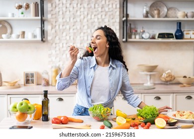 Young woman eating salad at table with organic vegetables, enjoying healthy diet, standing in light kitchen interior. Lady cooked veggie meal at home. Weight loss concept - Shutterstock ID 2159675181