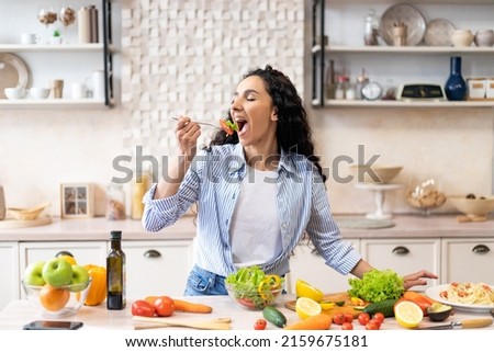 Young woman eating salad at table with organic vegetables, enjoying healthy diet, standing in light kitchen interior. Lady cooked veggie meal at home. Weight loss concept