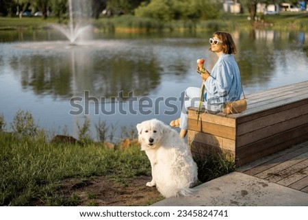 Young woman eats ice cream while sitting with her cute white dog near the lake in park. Friendship with pets and spending leisure time together Foto stock © 