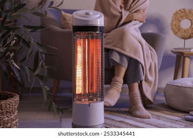 Young woman with electric heater and blanket sitting on armchair in evening at home, closeup