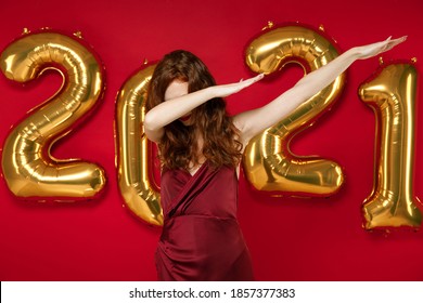 Young Woman In Elegant Dress Doing Dab Hip Hop Dance Hands Gesture, Youth Sign Hiding Isolated On Red Background Golden Numbers Air Balloons. Happy New Year 2021 Celebration Holiday Party Concept