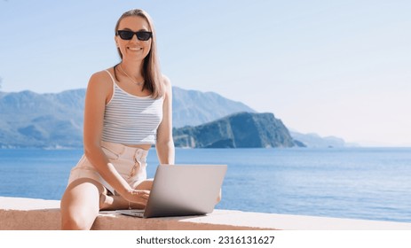 Young woman embraces the freelance lifestyle, effortlessly working on her laptop while appreciating the calming presence of the sea - Shutterstock ID 2316131627