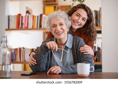 Young Woman Embracing Senior Mother At Home And Looking At Camera. Portrait Of Happy Adult Granddaughter And Grandmother Embracing And Smiling Together. Lovely Young Woman Hugging From Behind Grandma 