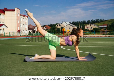 A young woman is engaged in fitness on the football field. Blue sky and bright sun. A sporty girl does gymnastic exercises on a mat.A healthy lifestyle.