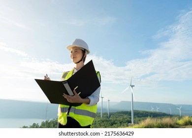 Young Woman Engineer Working With Report In Clipboard Against Wind Turbine Farm