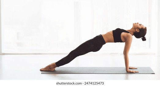 A young woman enhancing her core strength and balance through the reverse plank pose on a yoga mat. The spacious, sunlit studio sets a peaceful ambience for her morning workout routine