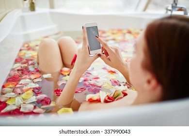 Young woman enjoying a bath with rose petals and using her phone - Powered by Shutterstock