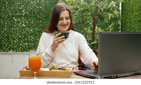Young woman enjoying breakfast on the terrace while working on her laptop. Concept of freelancing, working remotely, and having a meal outdoors - Shutterstock ID 2311237511