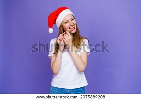 Young woman enjoying candy in santa hat and white t-shirt on lilac background. Girl chewing on a christmas lollipop and looking at the camera
