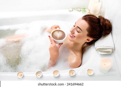 Young Woman Enjoying Coffee While Lying In The Bath With Foam And Candles