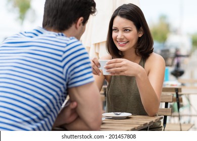 Young woman enjoying a cup of coffee outdoors at a street cafe as she sits chatting to her boyfriend or husband in the summer sun