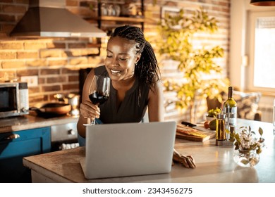 Young woman enjoying a glass of wine in the kitchen while on a video call on her laptop - Powered by Shutterstock