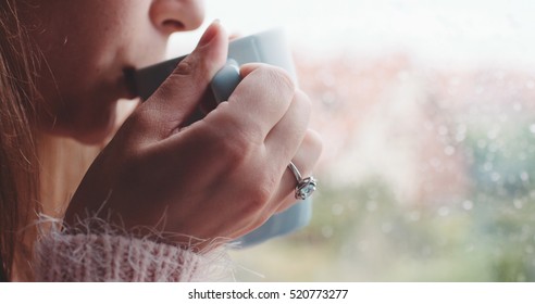 Young Woman Enjoying her morning coffee or tea, Looking Out the Rainy Window. Beautiful romantic unrecognizable girl drinking hot beverage at cozy home. Rainy Day Mood.