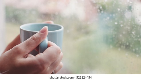 Young Woman Enjoying her morning coffee or tea, Looking Out the Rainy Window. Beautiful romantic unrecognizable girl drinking hot beverage at cozy home. Rainy Day Mood.