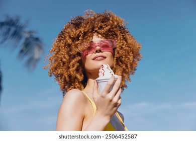 Young woman enjoying ice cream on a sunny day with a clear blue sky She has curly hair and wears stylish sunglasses - Powered by Shutterstock