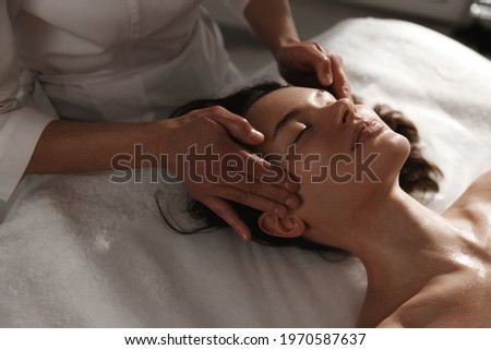 Young woman enjoying massage at spa salon. Beautician rubbing temples of client, smiling girl relaxing during myofascial massaging therapy at beauty clinic, cosmetology procedure
