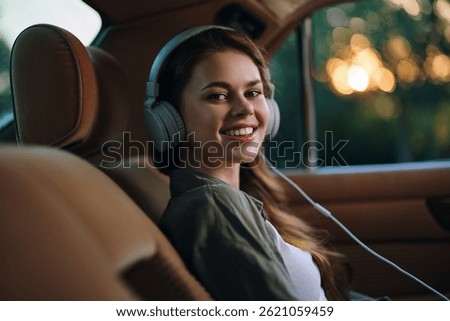 Young woman enjoying music in a car, smiling, wearing headphones, with a warm sunset background showcasing a relaxed and joyful mood.