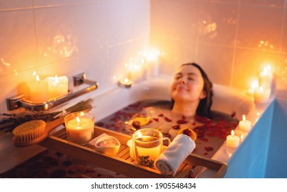 Young woman enjoying spiritual aura cleansing rose flower bath with rose petals and candles during full moon ritual. Body care and mental health routine. 