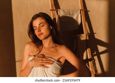 A young woman is enjoying the sun. A beautiful female model wrapped in a white towel stands in the bathroom after a shower. Shadows from tropical leaves. - Powered by Shutterstock