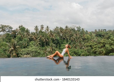 Young Woman Enjoying The Sun At Infinity Summer Swimming Pool At Luxurious Resort, Sit On The Edge, Jungle Forest On Background