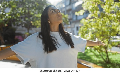 Young woman enjoying a sunny day in an urban park with arms outstretched, surrounded by greenery and city buildings in the background - Powered by Shutterstock