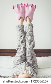 Young Woman Enjoying Sunny Morning. Caucasian Girl In Cute Warm Pajamas And Pink Monster Foot Slippers With Big Claws Lying On The Bed At Home Bedroom After Waking Up. Good Start Of The Day Concept.