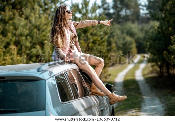 Young woman enjoying the\
trip while sitting on the car roof on a picturesque road in the\
woods