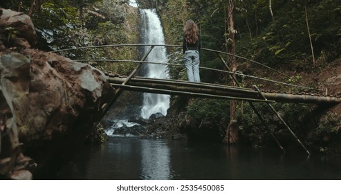 Young woman is enjoying the view of a waterfall in a tropical rainforest while standing on a bamboo bridge. She is surrounded by lush greenery and the sound of cascading water - Powered by Shutterstock