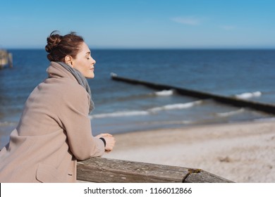 Young woman enjoying the warm sunshine at the seaside leaning on a wooden railing alongside the sea with closed eyes and a serene expression - Powered by Shutterstock