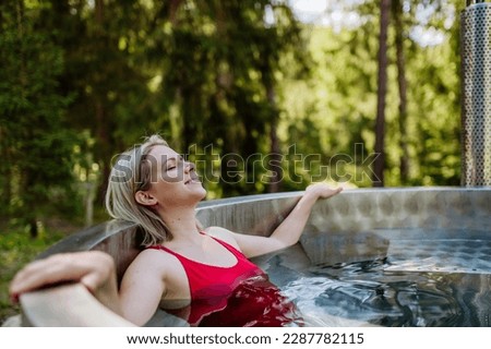 Young woman enjoying wooden bathtub with a fireplace to burn wood and heat water in backyard in mountains.