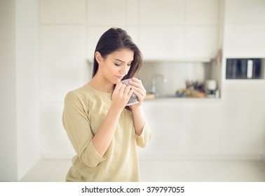 Young Woman Enjoying,holding Cup Of Hot Beverage,coffee Or Tea In Morning Sunlight.Enjoying Her Morning Coffee In The Kitchen.Savoring A Cup Of Coffee Breathing In The Aroma In Bliss And Appreciation