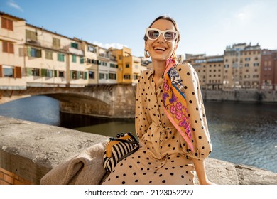 Young woman enjoys beautiful view on famous Old bridge in Florence, sitting on the riverside at sunset. Female traveler visiting italian landmarks. Stylish woman wearing dress and colorful shawl