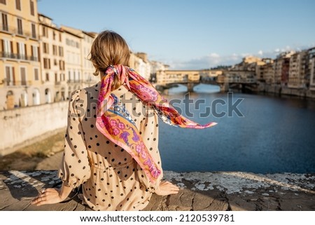 Young woman enjoys beautiful view on famous Old bridge in Florence, sitting back on the riverside at sunset. Female traveler visiting italian landmarks. Stylish woman wearing dress and colorful shawl