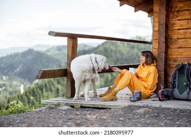 Young Woman Enjoys Great Mountain Landscape And Eats Sublimated Food For Hiking, While Sitting With Her Dog On A Wooden Terrace Of Nature House. Concept Of Solitude In Nature And Traveling With Pets