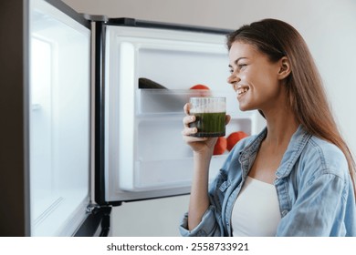 Young woman enjoys a healthy green smoothie while looking into the fridge, promoting healthy eating and a balanced lifestyle with fresh produce - Powered by Shutterstock