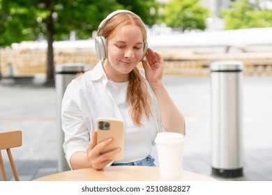 Young Woman Enjoys Music on Her Phone While Sipping Coffee in Outdoor Café Setting - Powered by Shutterstock