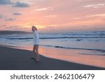 Young woman enjoys a peaceful walk along the shoreline as the sun sets, casting a soft glow across the sandy beach and gentle ocean waves
