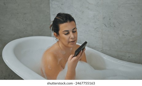 A young woman enjoys a relaxing bubble bath at home while browsing her smartphone in a contemporary bathroom setting. - Powered by Shutterstock