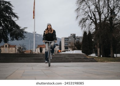 A young woman enjoys a solitary bike ride on a paved path in a serene urban park setting, with trees and city background. - Powered by Shutterstock