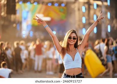 Young woman enjoys summer music fest, arms raised, peace sign, crowd, stage in background. Sunglasses, fun, excited fan at outdoor concert. Casual fashion, carefree youth at beach event.